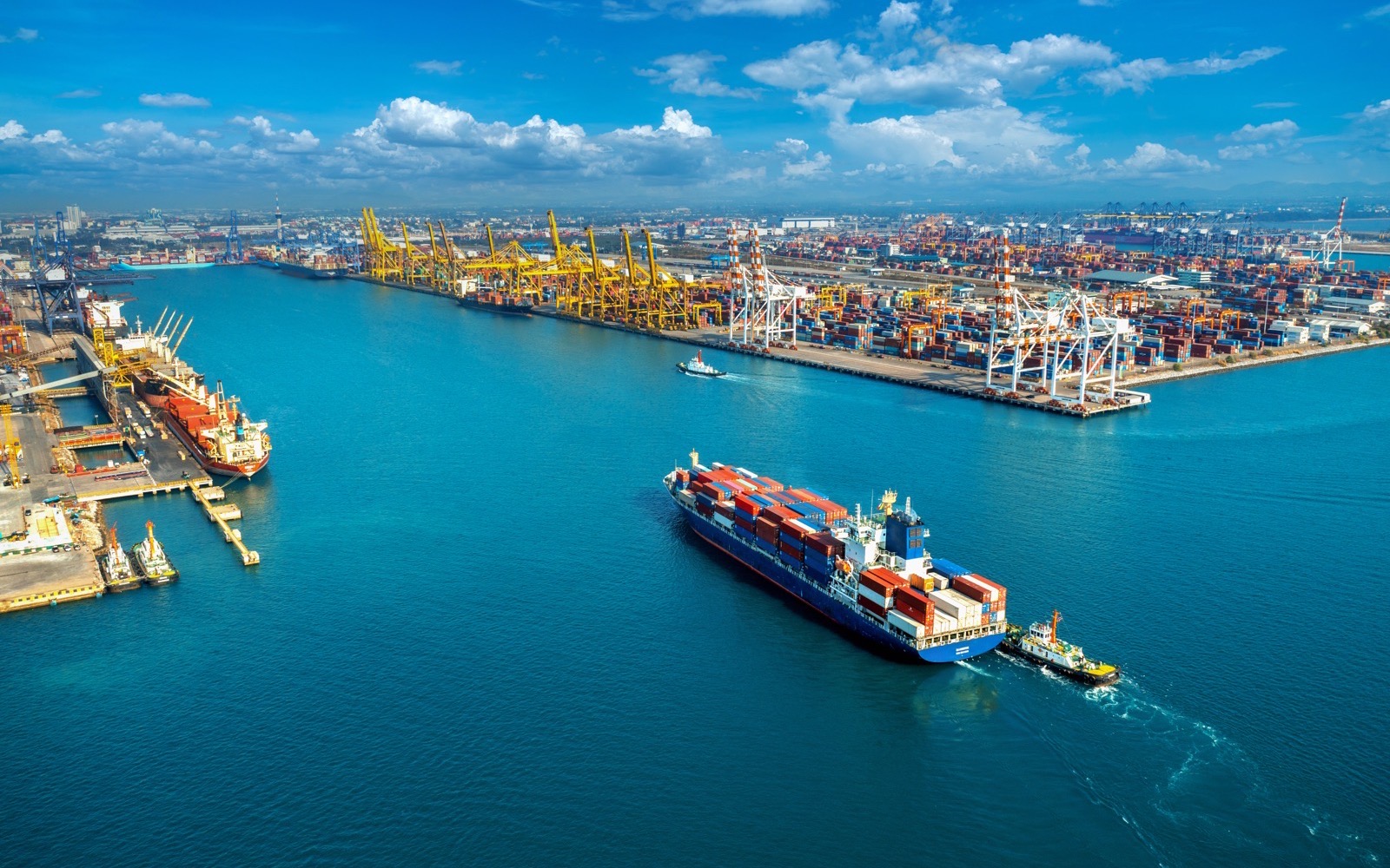 Container ship berthing at a port terminal, with gantry cranes and stacked containers along the quay